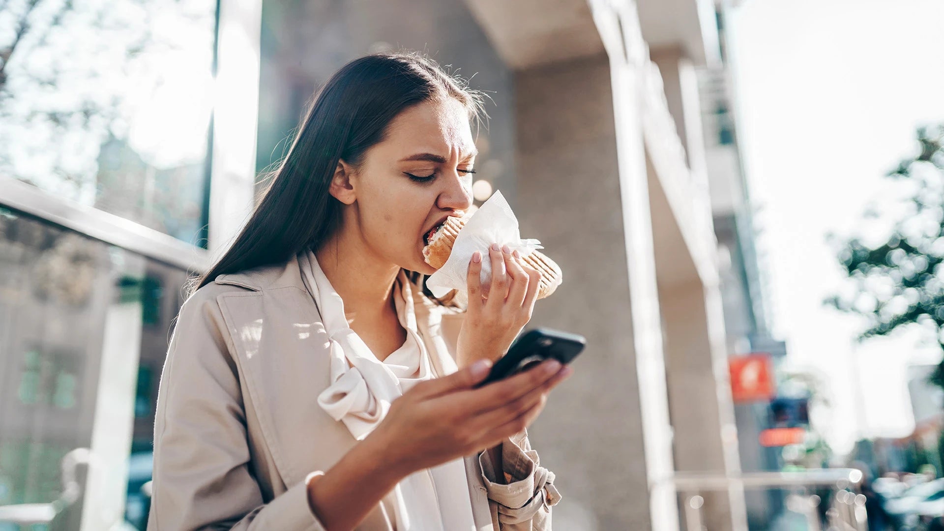 Young stressed woman eating a burger while checking her phone